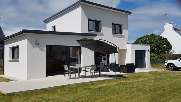 Uma casa moderna de dois andares com um espaço de varanda que inclui uma mesa, cadeiras e um grande guarda-sol, com um carro estacionado perto.