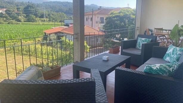 En balkon med fletningsmøbler, udsigt over et grønt marklandskab, og en bygning med rødt tag.