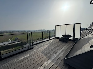 Une terrasse en bois dotée d’une balustrade en verre, offrant une vue sur un paysage rural.