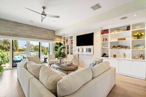 A modern living room with a large sofa, a coffee table, and a TV mounted on the wall. There are shelves with decorative items and a view of the outdoors. - Pass A Grille Don Cesar Pool Boat Slip 3/3.5 (St Pete Beach)