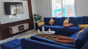 A living room with a blue sofa, a TV mounted on the wall, a plant, and a window with a view. - Djerba Aldo Piscine  (Houmt Souk)