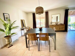 A dining area with a table, chairs, a hanging light fixture, a potted plant, a mirror, and a view of the outdoors.