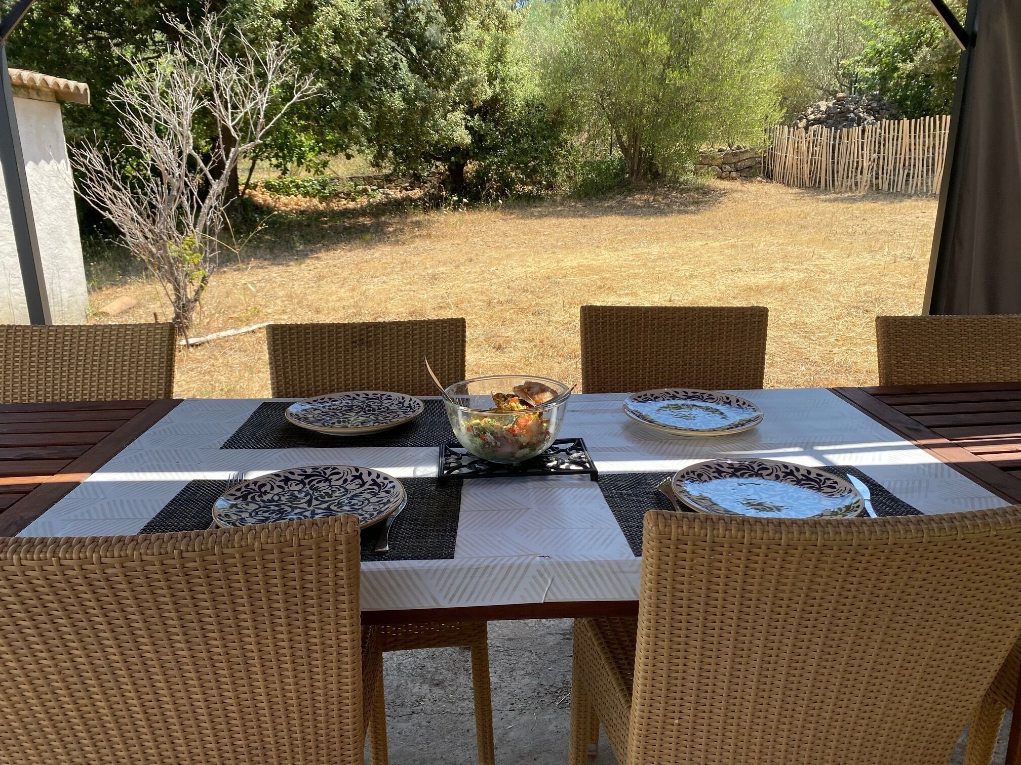 Un espace extérieur aménagé pour les repas, avec une table dressée pour quatre personnes, entourée de chaises en osier, et une vue sur un champ aride et des arbres en arrière-plan.