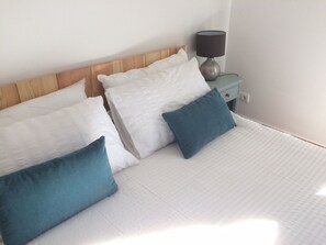 A neatly made bed with white bedding and two blue pillows, a wooden headboard, and a bedside table with a lamp.