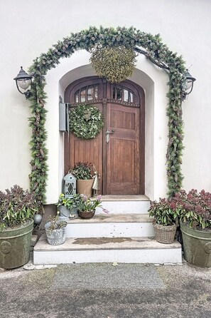 Une porte en bois ornée d’une guirlande, avec des plantes en pot et une lanterne posée sur les marches.