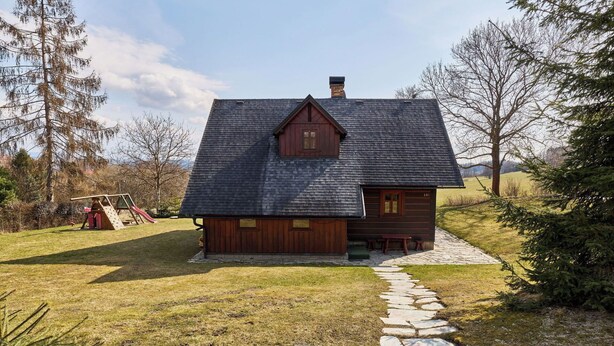 Un rustico chalet con un vialetto in pietra che conduce all'ingresso, immerso in un prato erboso e alberi.