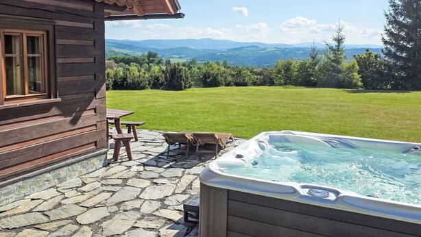 En træhytte med stenterrasse, jacuzzi og udsigt over en græsplæne og bjerge.