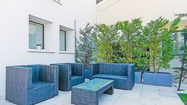 Uma área de estar moderna ao ar livre, com bancos fixos e uma pequena mesa, cercada por um edifício branco com grandes janelas e um arbusto verde.