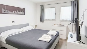 Quarto de hotel com uma cama, escrivaninha com computador, janela com cortinas e uma pintura com vista para a cidade na parede.
