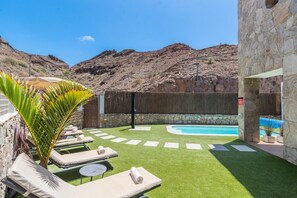A pool area with artificial grass, lounge chairs, and a stone wall. - Villa Chil - Luxury with heated pool and BBQ (Mogán)
