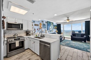 A modern kitchen with white cabinetry, a built-in microwave, and a dishwasher. There is a large window offering a view of the ocean. - FREE Golf! Boardwalk 2006 King Master, Walk in Shower, Hot Tubs, Heated Pool! (Panama City Beach)