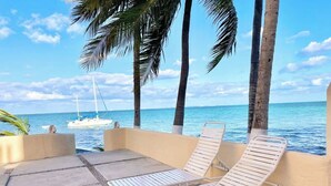 Eine Strandlounge mit zwei weißen Liegestühlen, einem Betonboden und Blick auf das Meer, im Hintergrund ein Segelboot.
