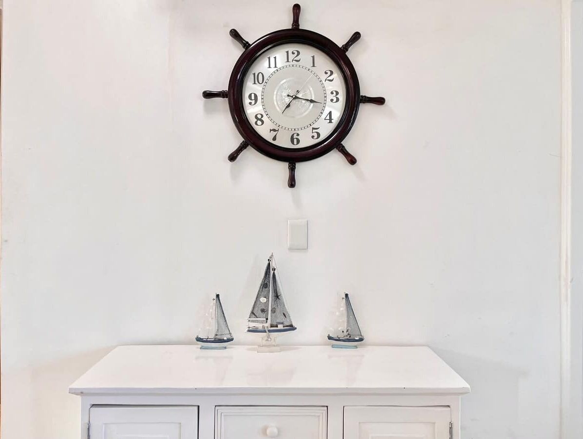 A wall-mounted clock with a ship's wheel design, a white dresser with three small sailboat decorations, and a white wall.