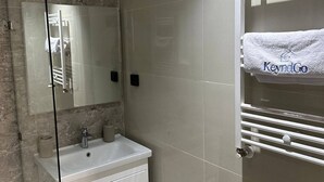 Ένα σύγχρονο μπάνιο με ένα διαχωριστικό ντους από γυαλί, έναν λευκό νιπτήρα και μια κρεμάστρα πετσετών με μια πετσέτα.