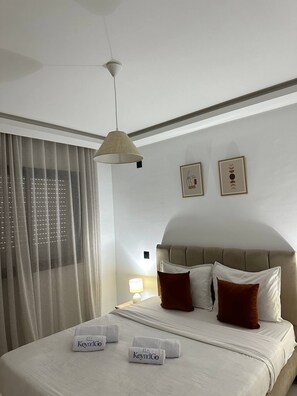 Habitación de hotel con cama, lámpara colgante, ventana con cortinas y dos cuadros en la pared.