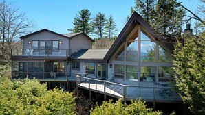 A modern two-story house with a deck, large windows, and a clear view of the surrounding forest. - Luxury Retreat w Gorgeous Lake Views, Minutes to Marina, Beach & Dining (Diamond Point)