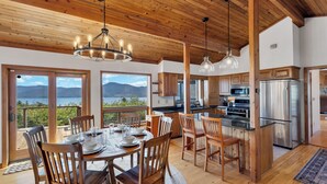A wooden interior with a dining table set for four, a kitchen with modern appliances, and large windows overlooking a scenic landscape. - Luxury Retreat w Gorgeous Lake Views, Minutes to Marina, Beach & Dining (Diamond Point)