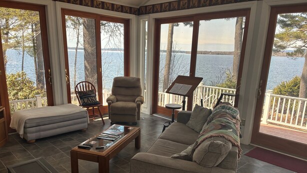 A spacious room with a large window overlooking a lake, a sofa, a chair, and a small table. - Spacious 5-bedroom lake house in Standish, ME (Standish)