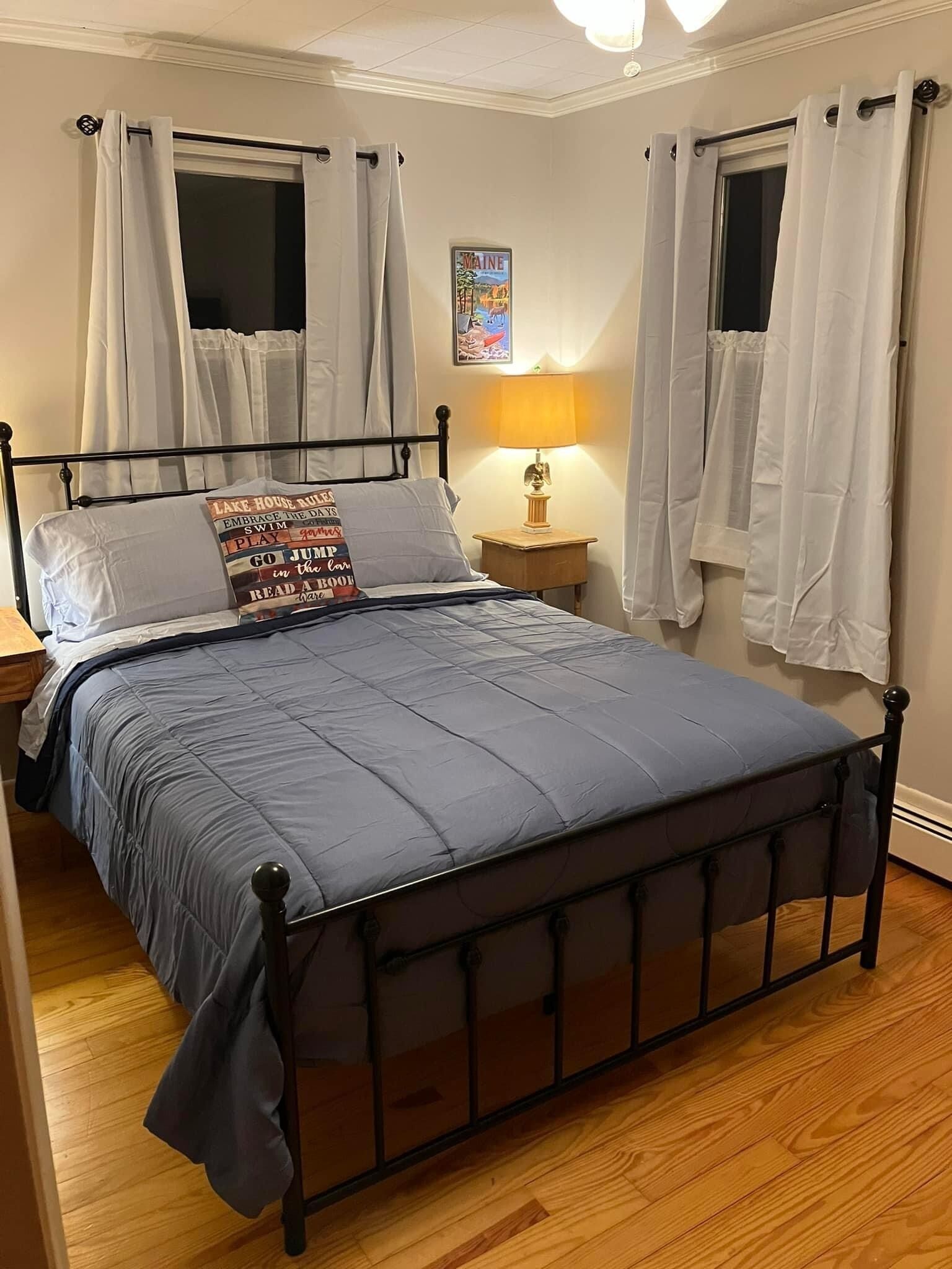 Une chambre à coucher avec un cadre de lit en métal, une couvre-lit bleue, des rideaux blancs, une petite table de chevet avec une lampe et un tableau accroché au mur.