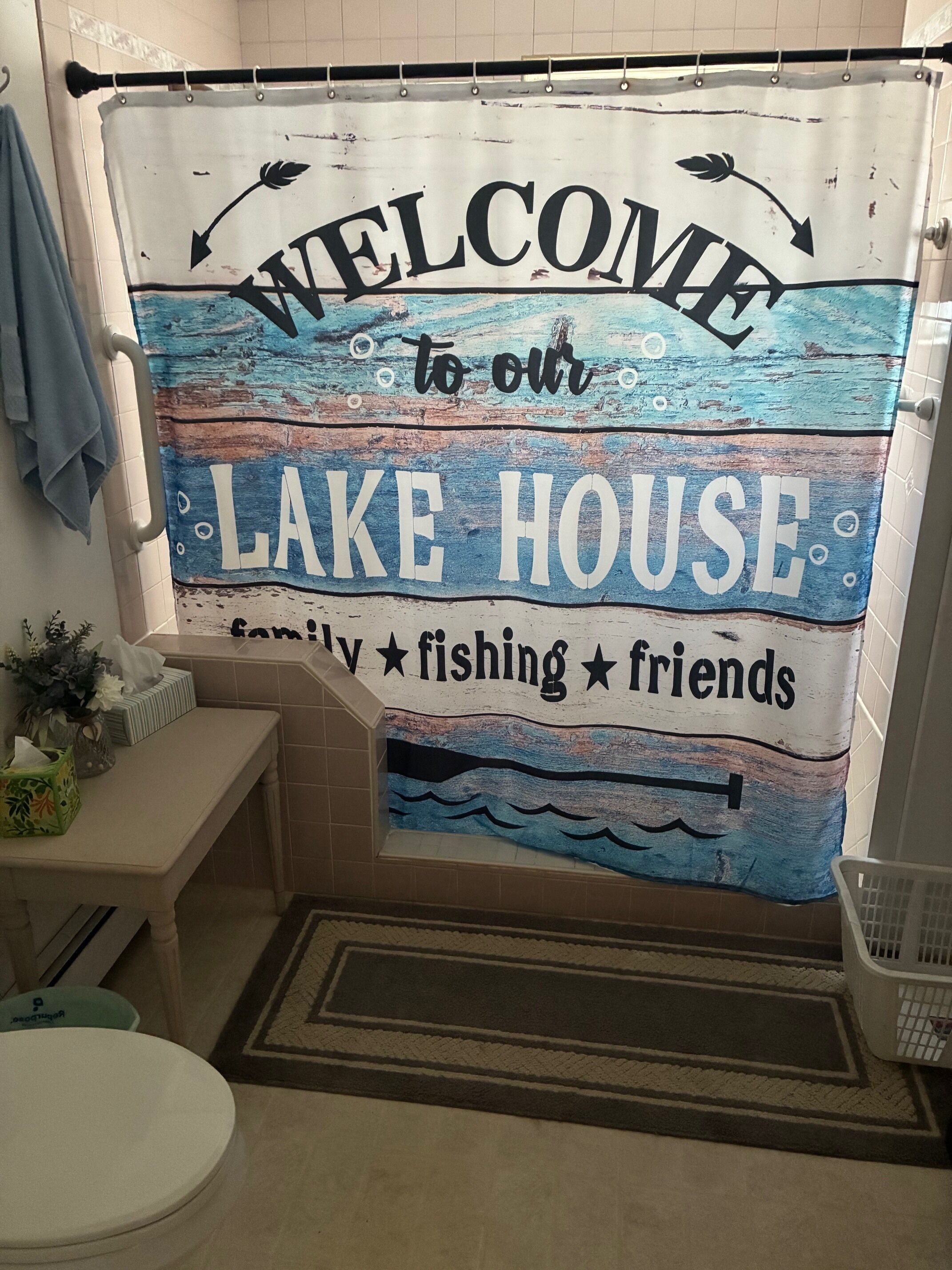 A bathroom with a 'Welcome to our Lake House' shower curtain, a toilet, a bench, and a small plant.