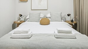 Un lit bien fait, avec du linge de lit blanc et un plaid à motifs, deux tables de chevet avec des lampes, et un tableau abstrait encadré accroché au mur.
