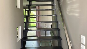 En moderne trappe med mørke trætrin og gelænder af glas, der fører op til en balkon med udsigt over grøn vegetation.