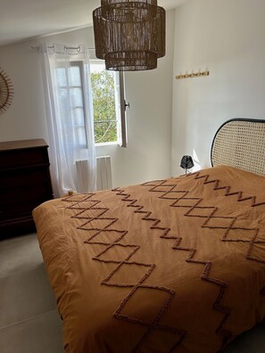 Un dormitorio con cama, cómoda, ventana con cortinas, lámpara colgante y aplique de pared.