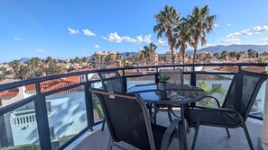 Un balcon avec une table et des chaises en verre, donnant sur une ville côtière bordée de palmiers et de bâtiments aux toits rouges.