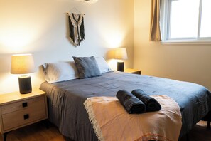 A bedroom with a bed, two bedside lamps, a nightstand, a window with curtains, and a wall-mounted tapestry. - cutest home in Sauble - 5 min. walk to the beach! (Sauble Beach)