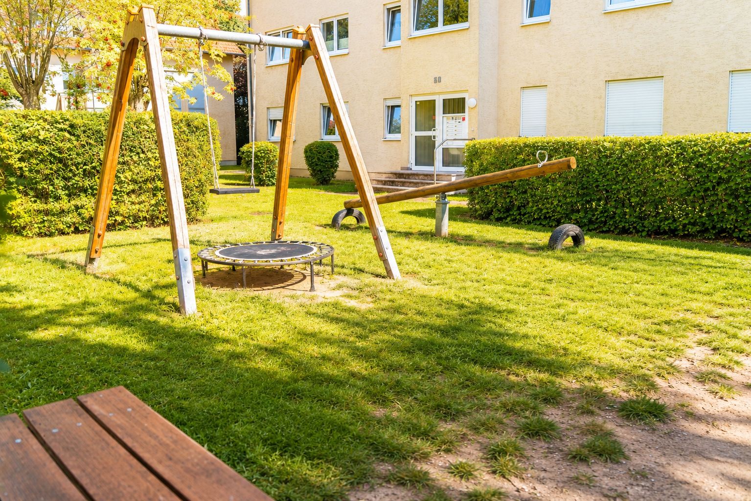 A playground with a swing set and a trampoline in a grassy area with a building in the background.