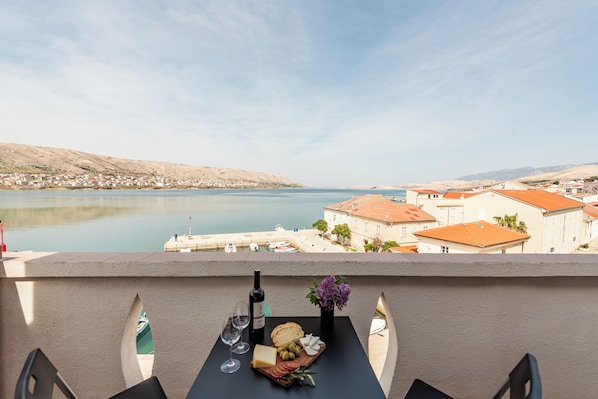 A balcony with a table set for a meal, overlooking a body of water and a coastal town.