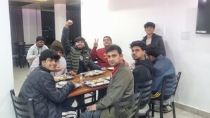 A group of people seated around a wooden table in a room with a white wall and a flat-screen TV mounted on it. - You home away from home in the mountains. (Shimla)