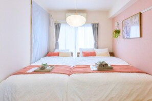 Ein Doppelbett mit weißer und pfirsichfarbener Bettdecke, eine hängende Pflanze und ein Bild an der Wand.