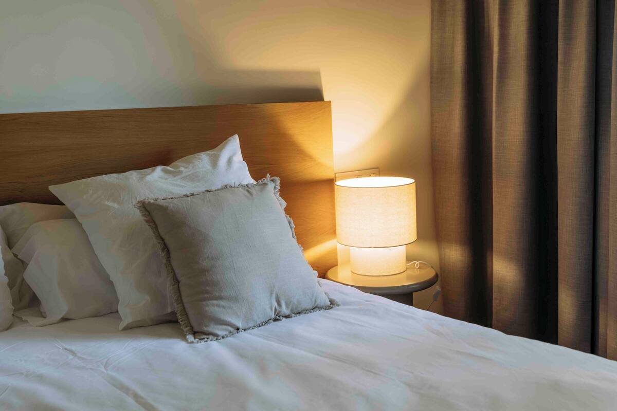 A neatly made bed with white linens and pillows, a bedside lamp, and a wooden headboard.