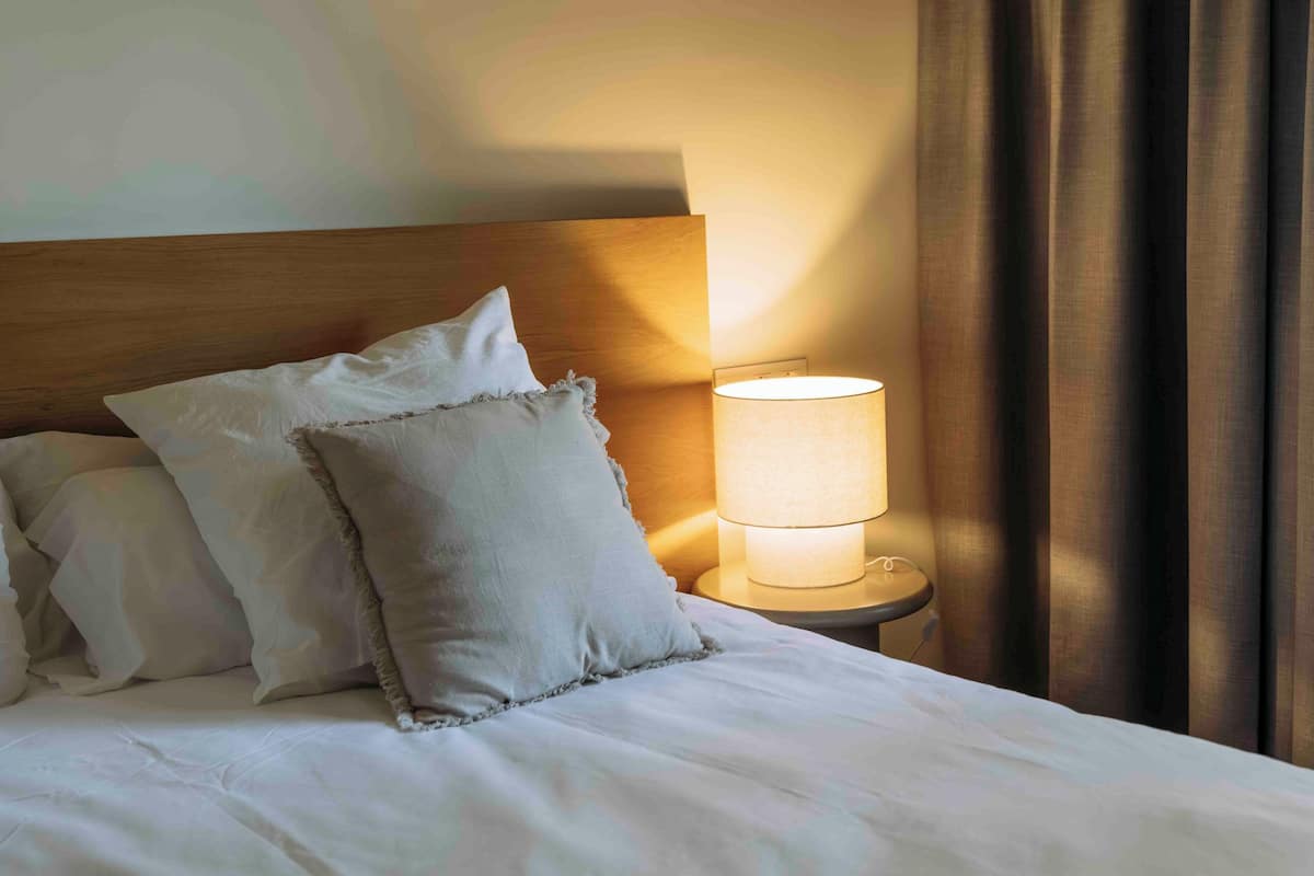 A neatly made bed with white linens and pillows, a bedside lamp, and a wooden headboard.