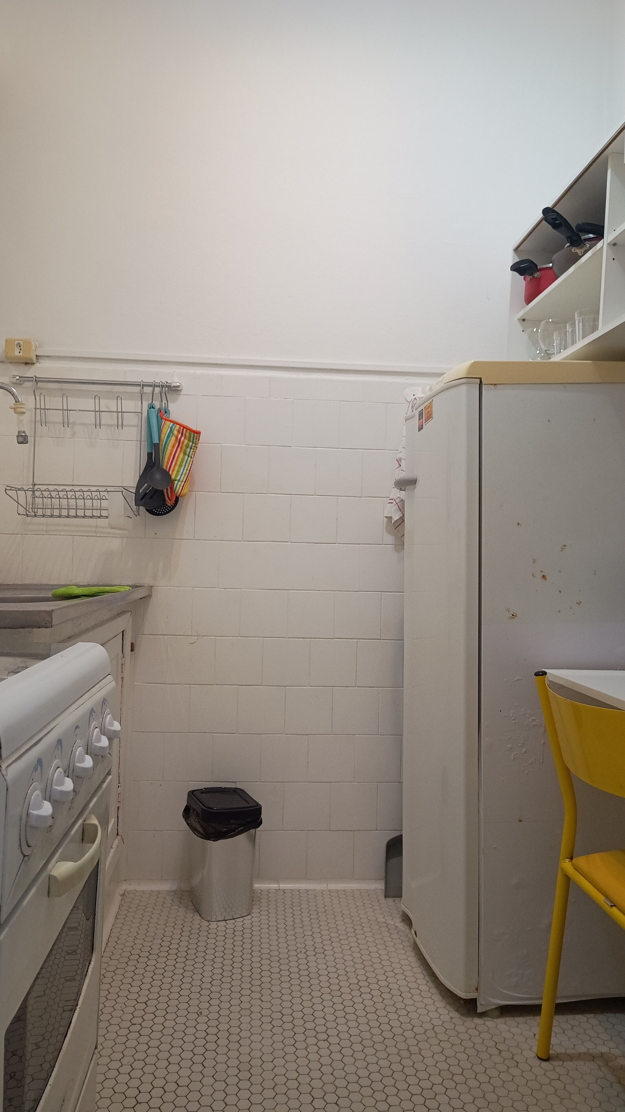 Une cuisine compacte avec des murs recouverts de carreaux blancs, une cuisinière blanche, une petite poubelle et une chaise jaune.