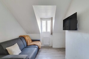A loft-style room with a gray sofa, a television mounted on the wall, and a window with curtains. - Charming Studio - 2P- le Marais (Paris)
