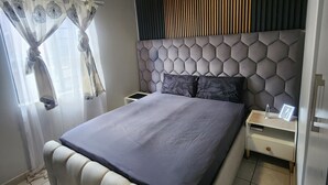 Une chambre à coucher moderne avec un grand lit, une table de chevet, une tête de lit ornée d’un motif en nid-d’abeille et un luminaire au plafond.