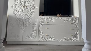 Ein Zimmer mit einem weißen Schrank mit aufwendigen Schnitzereien, einem Flachbildfernseher und Holzboden.