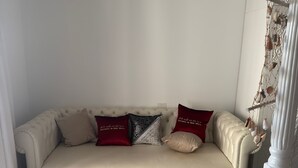 Ein beiges Sofa mit vielen Kissen, ein passender Hocker und eine Wanddeko mit Muscheln.