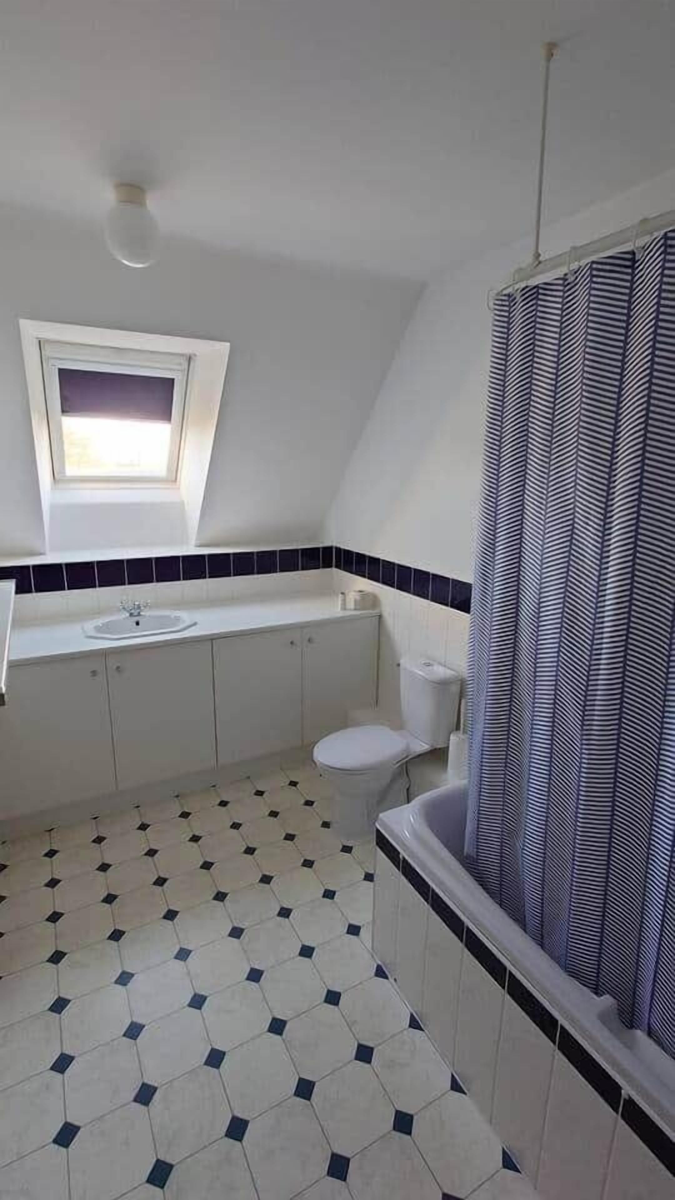 Ein Badezimmer mit weißem Waschbecken, Toilette und Badewanne, einem Fenster über dem Waschbecken und einem waschmusterigen Duschvorhang.