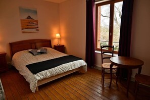 Ein Schlafzimmer mit Bett, Nachttischlampe, einem Fenster mit Vorhängen, einem Sessel und einem kleinen Tisch.