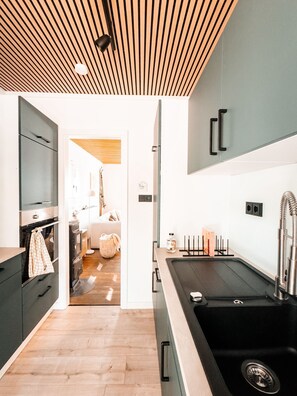 A modern kitchen with a wooden ceiling, a black sink, and a view into the adjacent living area.