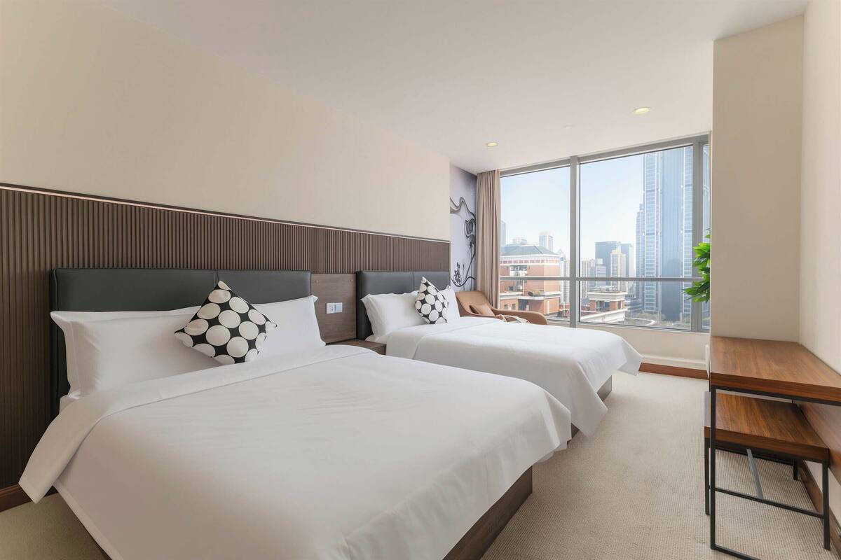 Une chambre d’hôtel avec deux lits, un bureau en bois et une grande fenêtre donnant sur la ville.