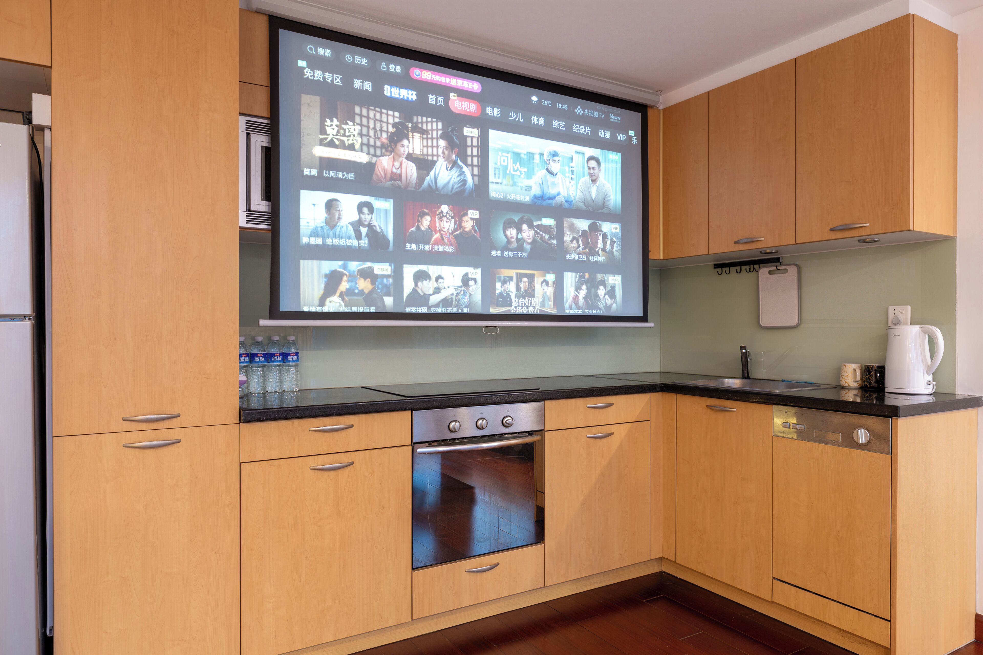 A kitchen with wooden cabinets, a built-in oven, a black countertop, and a large screen displaying various content.