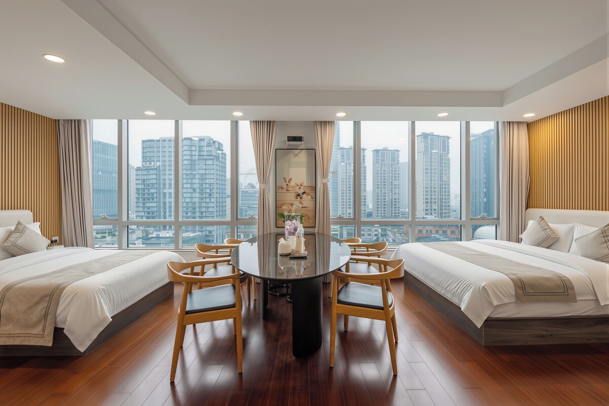 A hotel room with a large window, a round dining table, two beds, and wooden flooring.