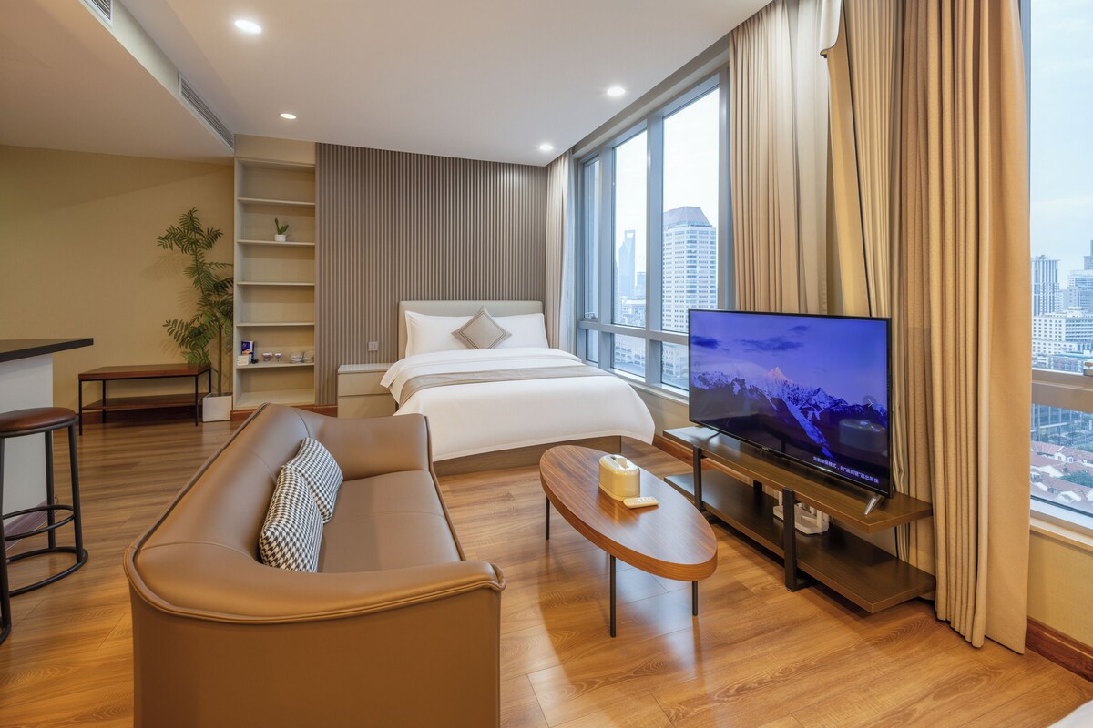 A modern hotel room with a sofa, a bed, a TV, and a city view.