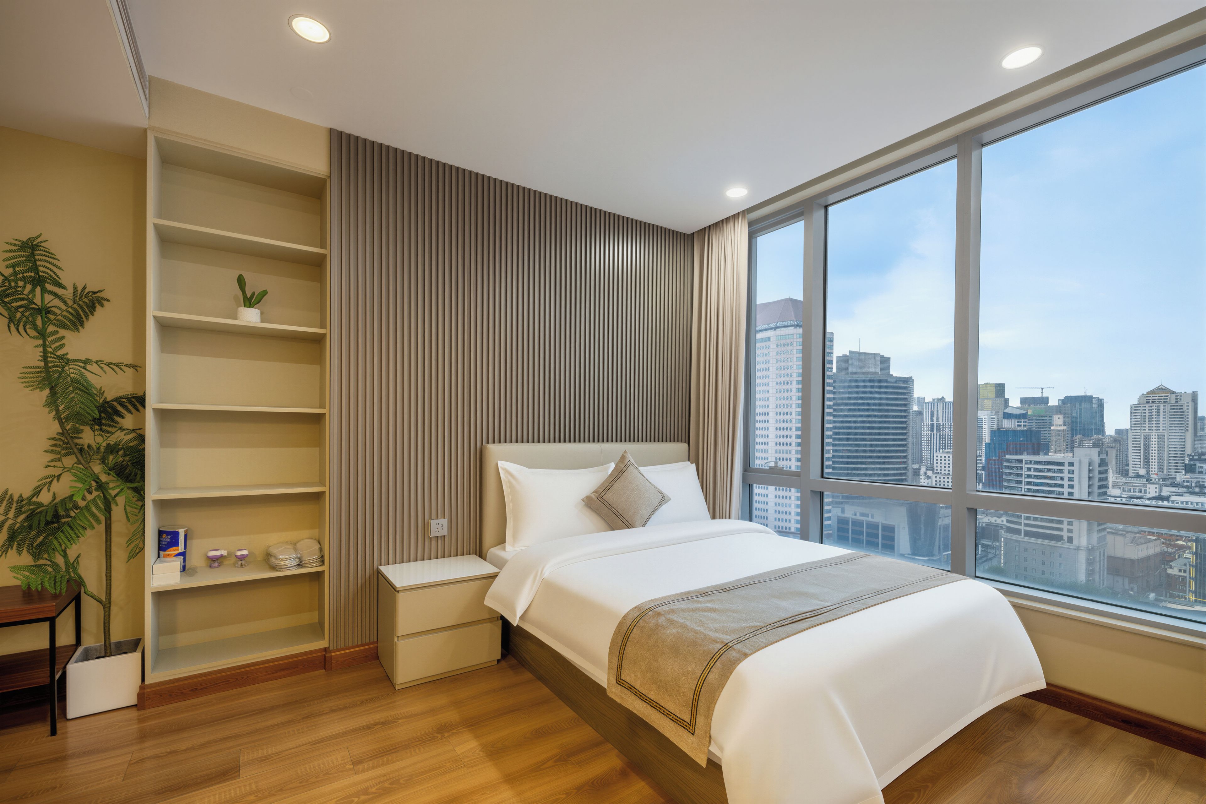 A modern hotel room with a large window, a bed, a bedside table, a shelf with a plant, and a view of the city skyline.