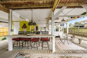 A covered outdoor dining area with a long table and bar stools, a hanging light fixture, and a wooden deck. - Walkable Family Beach Base, Pet Friendly (Gulf Shores)