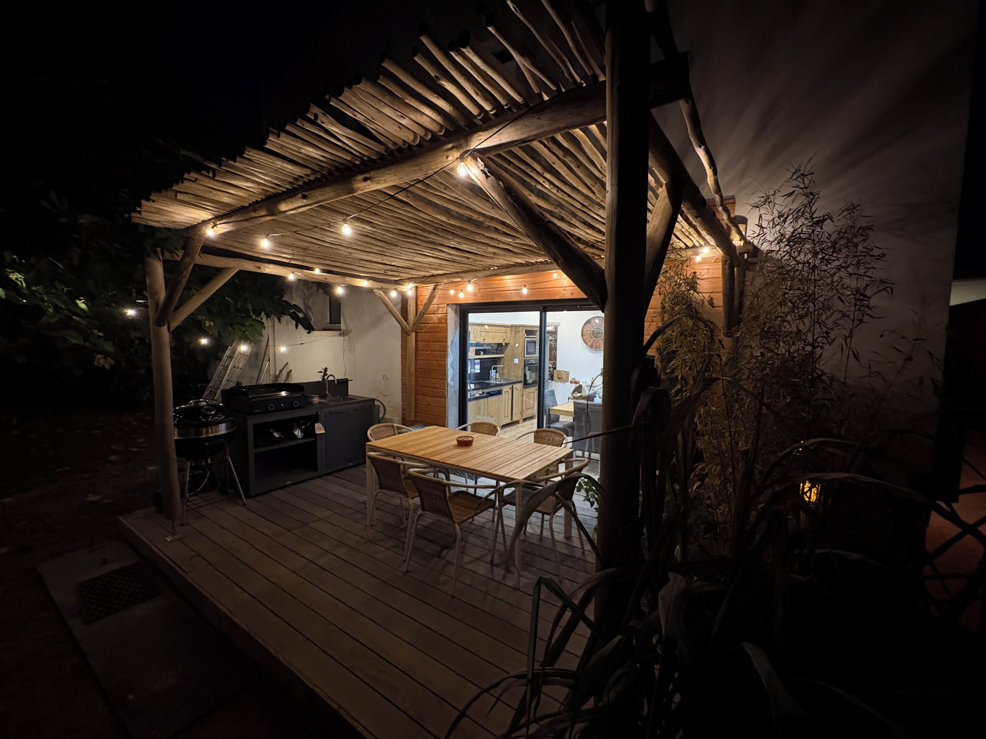Une terrasse extérieure avec une plateforme en bois, une table à manger avec des chaises, un barbecue et des guirlandes lumineuses.